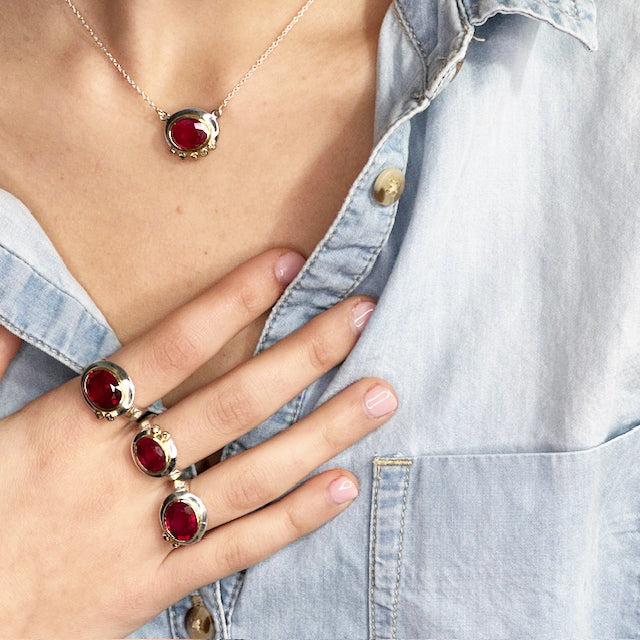 Person wearing a silver necklace with a red gemstone and multiple matching rings, against a denim shirt background.