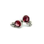 A pair of silver stud earrings featuring round deep pink tourmaline gemstones  with smaller round diamond accent stones clustered on the side, set against a plain white background.