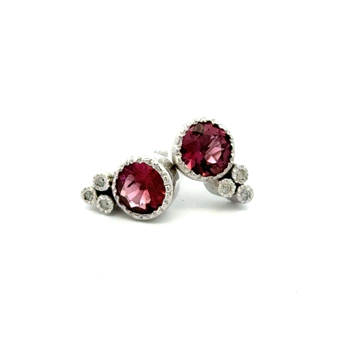 A pair of silver stud earrings featuring round deep pink tourmaline gemstones  with smaller round diamond accent stones clustered on the side, set against a plain white background.