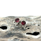 A pair of silver stud earrings with round deep pink tourmaline gemstones and small diamond accent stones, displayed on a textured piece of driftwood against a white background.