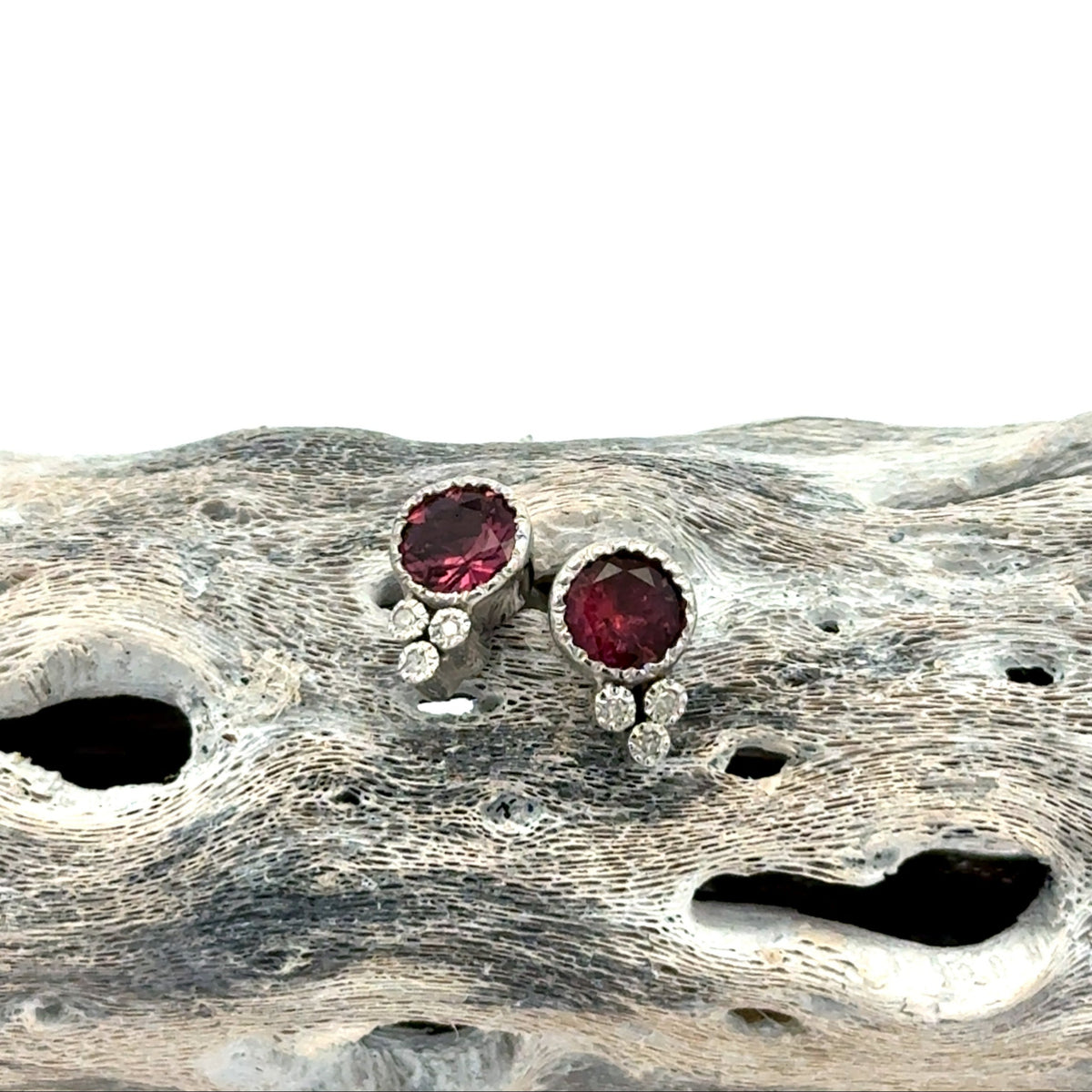 A pair of silver stud earrings with round deep pink tourmaline gemstones and small diamond accent stones, displayed on a textured piece of driftwood against a white background.