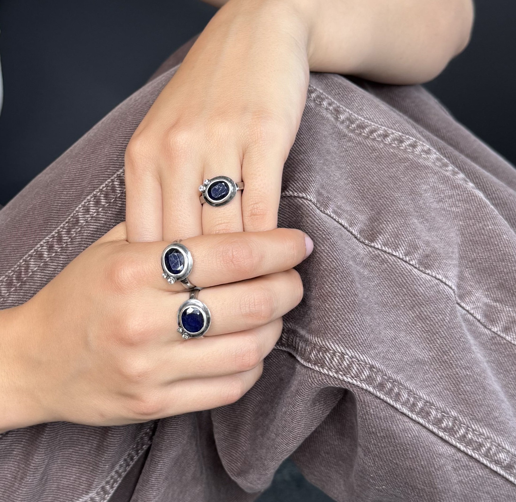 Close-up of hands wearing three silver rings with blue gemstones on a dark background